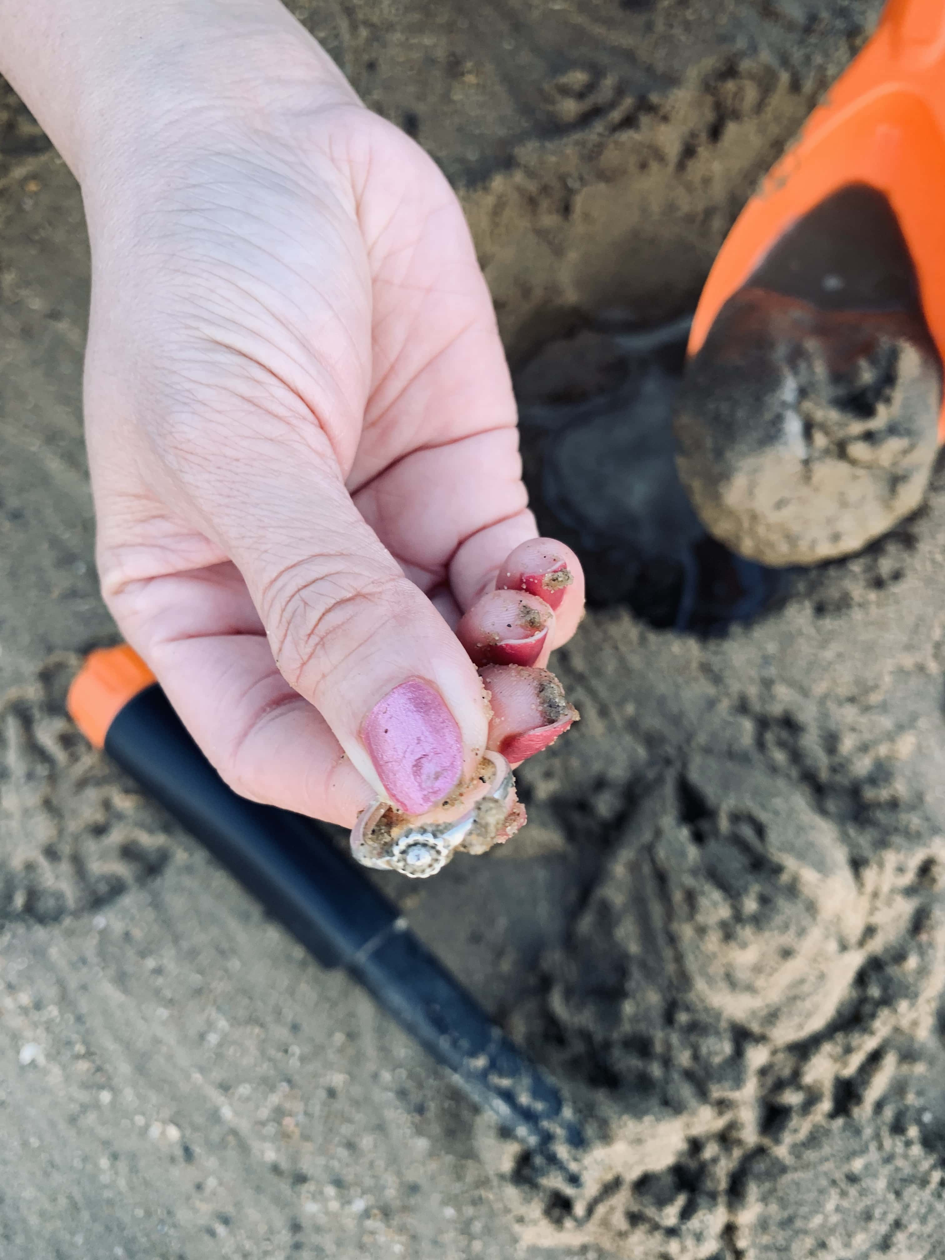 Hand Holding Ring with Starburst Gem Setting with Sandy Painted Nails and Pinpointer in Background on Sand Beach
