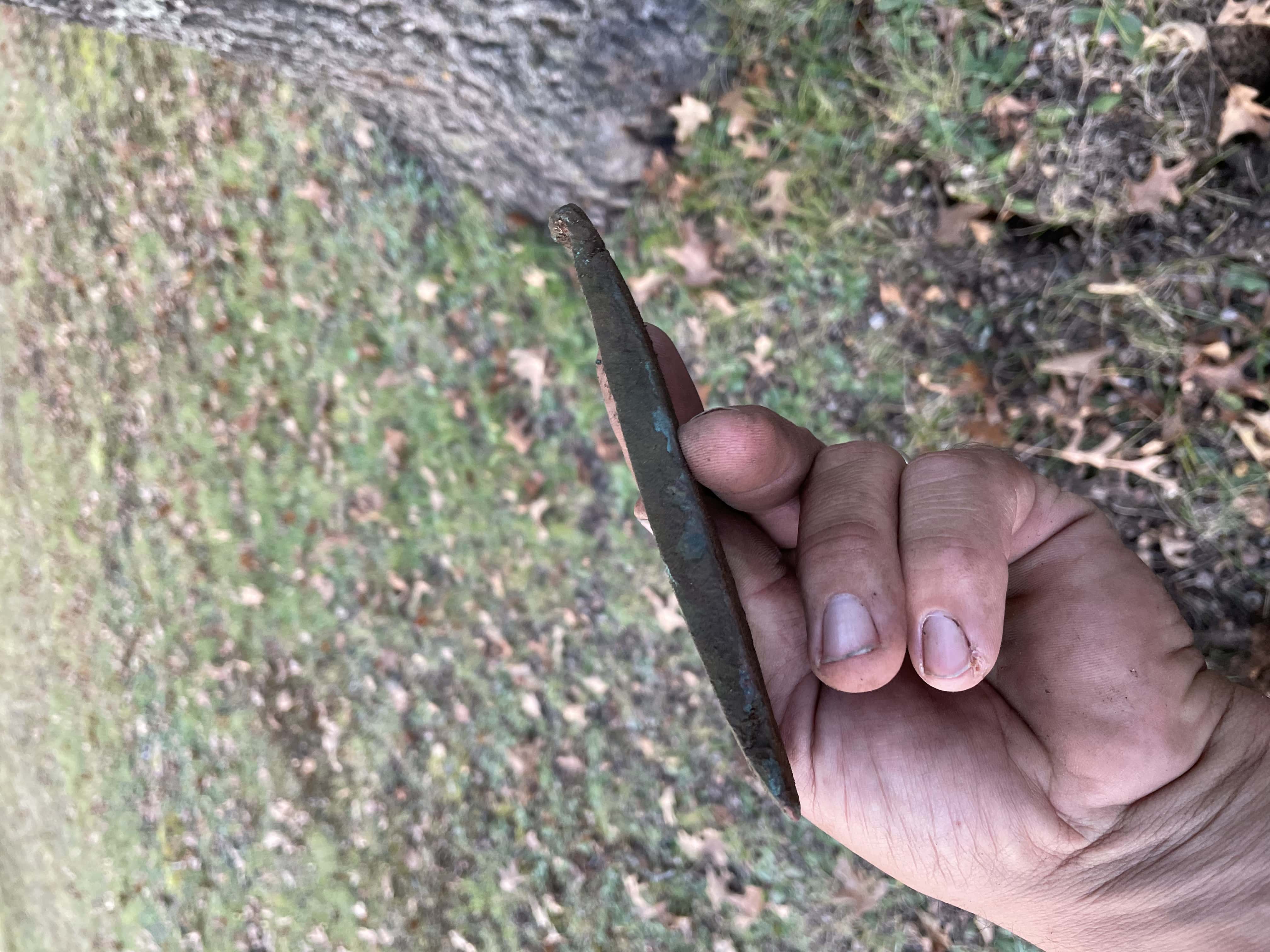 Prehistoric Copper Implement with Green Patina Across Surface Held in a Hand with Grassy Park and Tree Blurry in Background