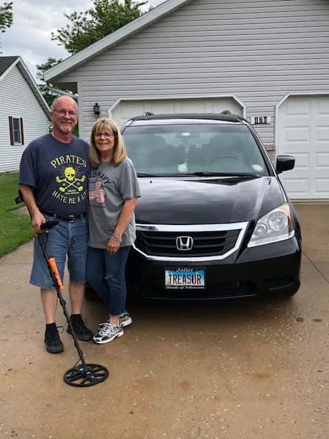 A Man Holding an Equinox 800 Metal Detector with a Garrett Carrot Pinpointer Strapped to it With Wife Standing In front of a Car that Reads