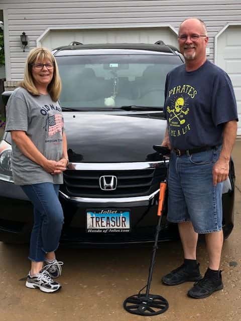 A Man Holding an Equinox 800 Metal Detector with a Garrett Carrot Pinpointer Strapped to it With Wife Standing In front of a Car that Reads